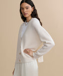 A woman with long dark hair wears the Cropped Cashmere Cocoon Cardigan over a white top and light pants, standing against a beige background. She accessorizes with gold jewelry and looks toward the camera with a neutral expression.