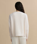 A person with long dark hair, seen from behind, wears the Cropped Cashmere Cocoon Cardigan in ivory with light pants, standing against a plain beige background.