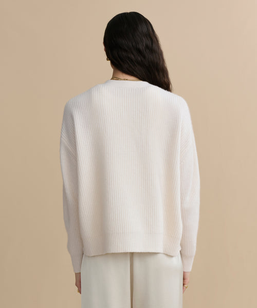 A person with long dark hair, seen from behind, wears the Cropped Cashmere Cocoon Cardigan in ivory with light pants, standing against a plain beige background.