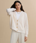 A woman with long dark hair wears the Cropped Cashmere Cocoon Cardigan in ivory over a white shirt and cream pants, accessorized with gold hoop earrings and layered necklaces, standing against a beige background.