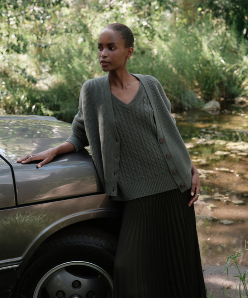 A woman in a Cropped Cashmere Cocoon Cardigan and pleated skirt leans against a car outdoors, surrounded by lush greenery and sunlight.