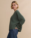 A woman with short blonde hair wears the Cropped Cashmere Cocoon Cardigan with blue jeans, standing sideways with one hand in her pocket against a plain beige background.