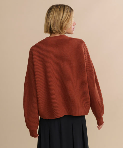 A person with short blonde hair wearing a Cropped Cashmere Cocoon Cardigan in rust and a black skirt stands against a beige background, facing away from the camera.