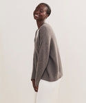A woman with short hair smiles while standing and tilting slightly forward. She wears the Cropped Cashmere Cocoon Cardigan paired with a white skirt against a plain light background.
