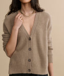 A woman wears the Cropped Cashmere Cocoon Cardigan with a deep V-neck, layered gold necklaces, and a gold ring, standing against a plain beige background. Her face is partially out of frame.
