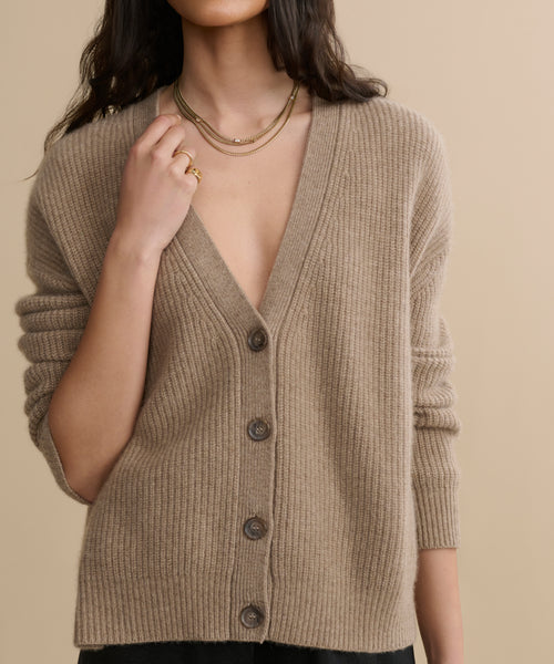 A woman wears the Cropped Cashmere Cocoon Cardigan with a deep V-neck, layered gold necklaces, and a gold ring, standing against a plain beige background. Her face is partially out of frame.