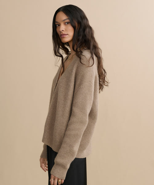 A woman with long, wavy brown hair wears the Cropped Cashmere Cocoon Cardigan in undyed stone and a black skirt, standing before a plain beige background and gazing slightly to the side with a neutral expression.