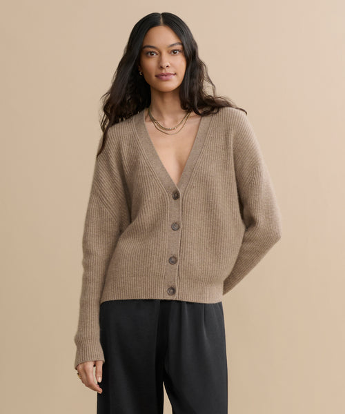 A woman with long dark hair wears the Cropped Cashmere Cocoon Cardigan in undyed stone with a deep V-neck and black pants, standing against a beige background.