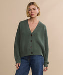 A person wears the Cropped Cashmere Cocoon Cardigan in olive green with a V-neck and blue jeans, standing against a plain beige background.