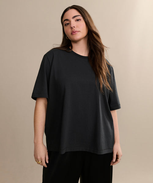 A woman with long brown hair wears the Crosby Tee, a relaxed fit shirt made from premium jersey cotton, paired with black pants. She stands against a plain beige background, looking at the camera with a neutral expression.