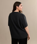 A woman with long brown hair wears the Crosby Tee, a relaxed fit top in premium jersey cotton, paired with black pants. She stands with her back turned, looking to the side against a plain beige background.