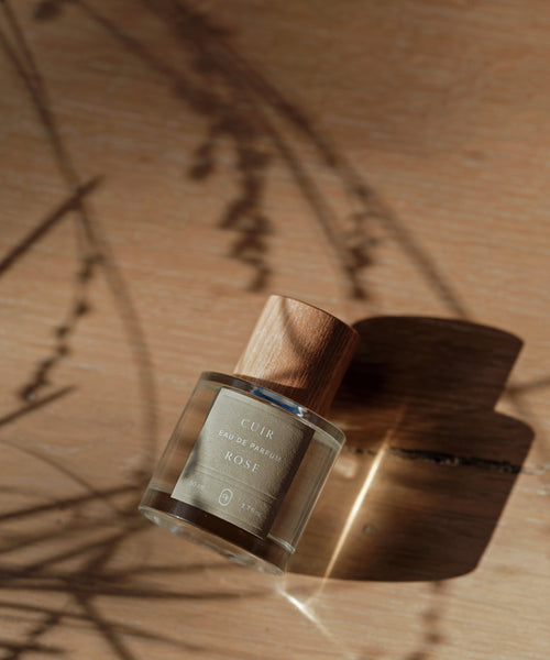 Cuir Rose Eau De Parfum, in a glass bottle with a wooden cap, rests on wood and is lit by sunlight and subtle plant shadows, reflecting the clean fragrance’s floral and woody notes.