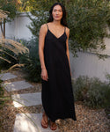 A woman with long dark hair stands outside on a stone path, surrounded by green plants and a white fence, wearing the Rio Slip Dress in black with brown sandals.