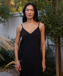 A woman with long dark hair stands outdoors in front of green foliage and a white fence, wearing the Rio Slip Dress—a sleeveless black dress with thin straps. She looks directly at the camera, her expression neutral.