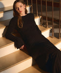 A woman with long brown hair wears the Dara Top, featuring black ribbed fabric and bell sleeves, as she sits on wooden stairs in warm natural light, calmly looking at the camera.