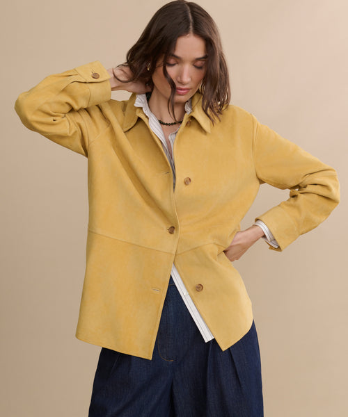 A woman poses against a beige background, wearing the Darcy Shirt—an ivory pinstripe top—layered under a loose yellow jacket with dark wide-leg pants. One hand is behind her head, the other on her hip.