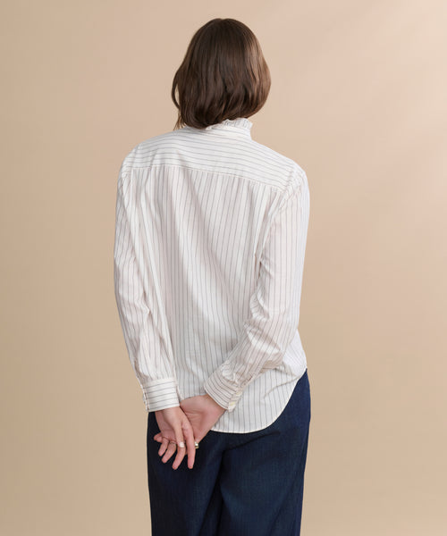 A person with short brown hair, wearing the ivory Darcy Shirt and dark pants, stands facing away with hands clasped behind their back against a plain beige background.