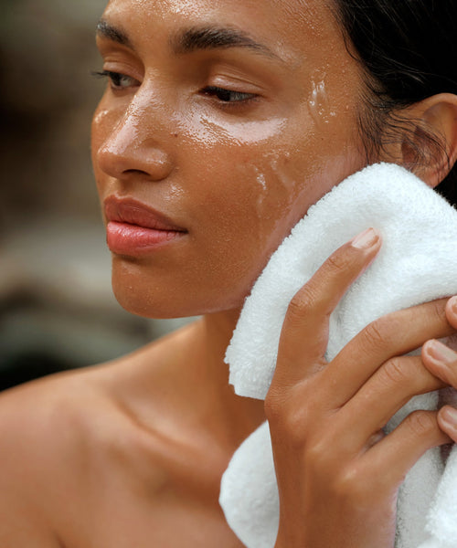 A woman gently presses a white towel to her cheek, her skin glistening with moisture after cleansing with Cleansing Balm.