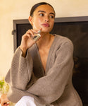 A woman with pulled-back hair and a loose beige sweater holds a Cuir Rose Eau De Parfum bottle near her neck, sitting indoors and gazing thoughtfully, evoking the clean floral and woody notes of this fragrance.