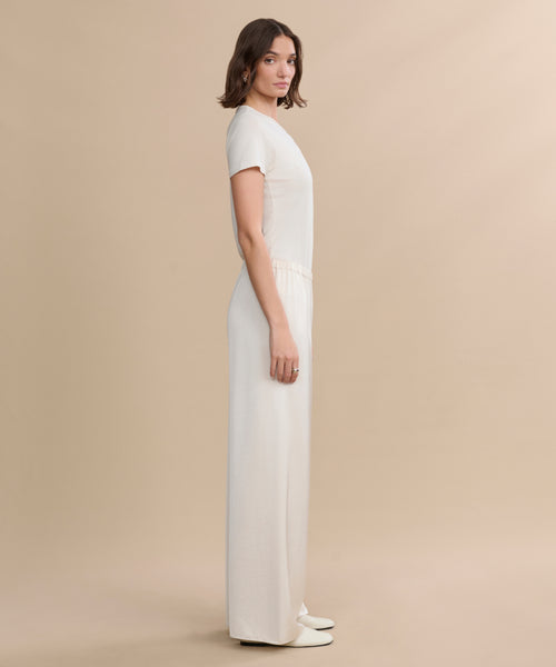 A woman with shoulder-length brown hair and a neutral expression stands sideways against a beige background, wearing the cream-colored Demi Pant and white shoes. The outfit has a flowy, short-sleeved look.