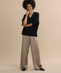 A woman stands against a beige background, smiling as she touches her face while wearing wide-legged taupe satin Demi Pants and a loose black V-neck sweater for an effortlessly flowy look.