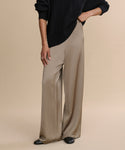 Model poses against a beige backdrop, showcasing the high-waisted, flowy Demi Pant in taupe satin, styled with a black ribbed sweater and black shoes. Only the lower half of the look is visible.