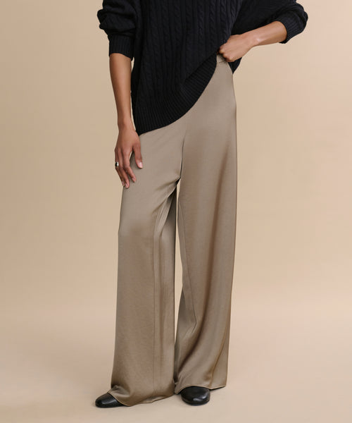 Model poses against a beige backdrop, showcasing the high-waisted, flowy Demi Pant in taupe satin, styled with a black ribbed sweater and black shoes. Only the lower half of the look is visible.