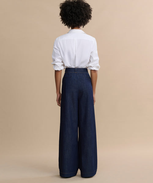 A person stands facing away, wearing a white long-sleeve shirt with rolled-up sleeves and Denim Newport Trousers featuring a high-waisted, straight-leg fit, set against a plain beige background.