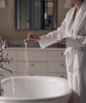 A person in a white bathrobe adds Relaxation Bath Set soak to a freestanding tub with running water in a softly lit bathroom. The persons face is not shown.