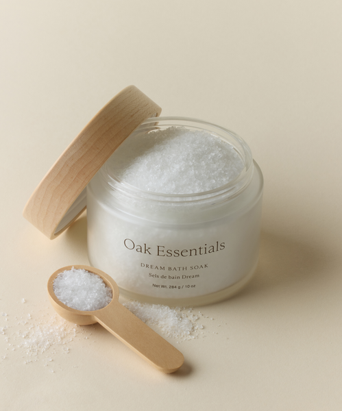 A glass jar of Dream Bath Soak, crafted with European sea salt and a wooden lid, rests beside a small wooden spoon filled with white bath salts—perfect for your pre-sleep ritual—on a light beige surface.
