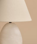 A close-up of the Droplet Alabaster Table Lamp featuring a beige linen shade and rounded, marble-like base, set against a neutral beige background.