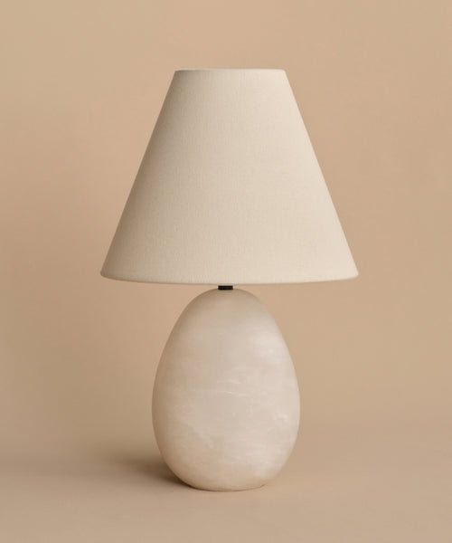 The Droplet Alabaster Table Lamp features an oval stone-like base and a wide linen shade, making it a stylish choice for bedside use against a plain beige background.