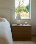 A minimalist bedside table with two drawers holds a Droplet Alabaster Table Lamp with a linen shade, a lit candle, and a small dish. Soft natural light from the window behind brightens a bed with light bedding on the left.