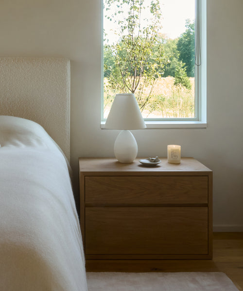 A minimalist bedside table with two drawers holds a Droplet Alabaster Table Lamp with a linen shade, a lit candle, and a small dish. Soft natural light from the window behind brightens a bed with light bedding on the left.