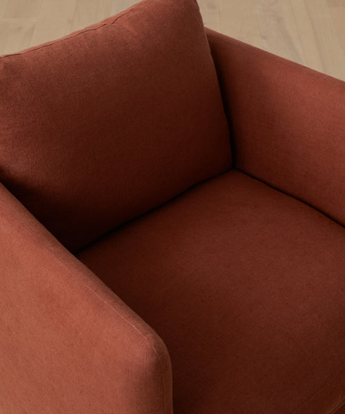 A close-up of the Dume Chair, a sustainable linen chair with rust-colored upholstery, highlighting its seat cushion, backrest, and armrests on a light wooden floor.