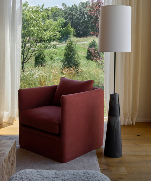 A stylish Dume Chair with a hypoallergenic pillow sits beside a tall black floor lamp by large windows, offering views of green trees and sunlight streaming in.
