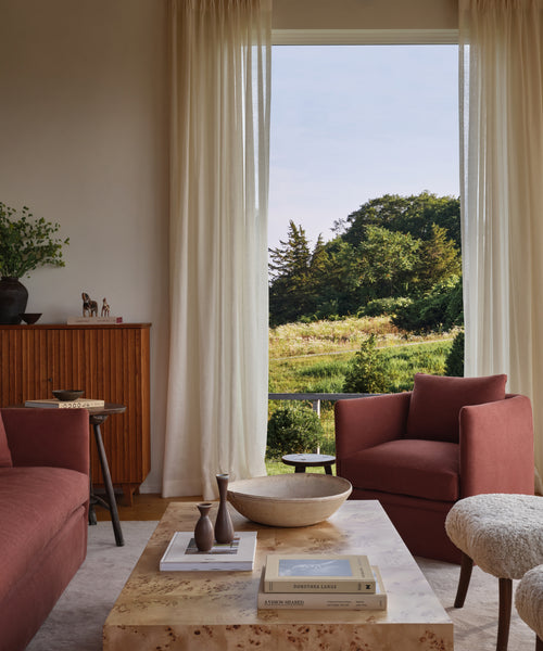 A cozy living room featuring the Dume Chair in rust, a marble coffee table with books and vases, a hypoallergenic pillow for comfort, and a large window with sheer curtains framing a green, sunlit landscape.