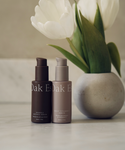 Two bottles of The Serums by Oak Essentials—one dark brown, one taupe—sit on a marble surface in front of white tulips, a chic duo to elevate your skincare routine.