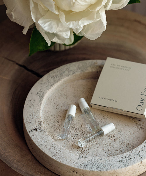 The Eau De Parfum Discovery Set, featuring three small perfume sample vials in a cream-colored box, sits on a round stone tray beside a white flower with green leaves, all arranged on a wooden surface.
