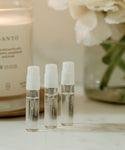 Three Eau De Parfum Discovery Set sample spray bottles stand upright on a white surface, with a candle and vase of white flowers blurred in the background—a luxe way to explore signature scents.