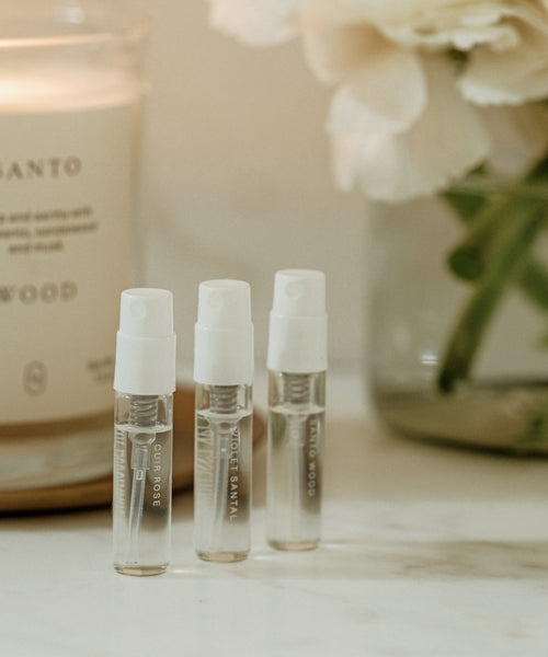 Three Eau De Parfum Discovery Set sample spray bottles stand upright on a white surface, with a candle and vase of white flowers blurred in the background—a luxe way to explore signature scents.