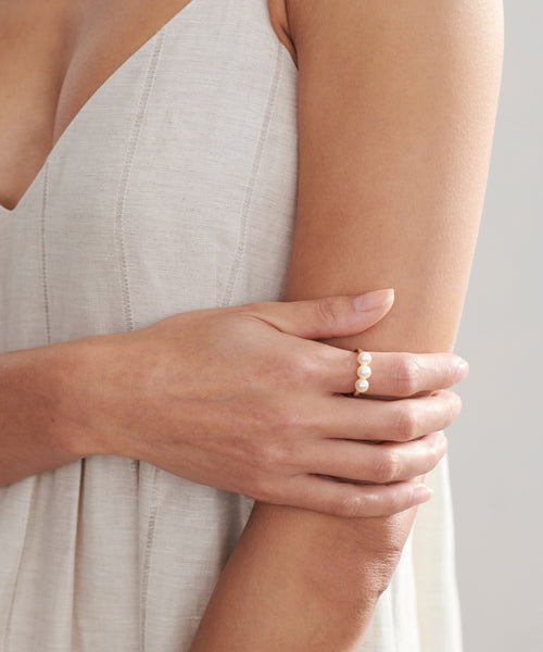 A person in a sleeveless, light beige dress showcases the Elodie Pearl Ring, a 14k gold ring set with four pearls, against a softly lit neutral background that highlights its elegant design.