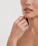 A woman with natural makeup wears a gold necklace with a single pearl pendant and the Elodie Pearl Ring, a 14k gold ring adorned with three pearls, gently touching her chin against a light background.