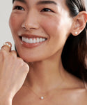 A woman smiles, showing white teeth. She wears pearl stud earrings, a delicate gold necklace with a single pearl, and the Elodie Pearl Ring—a 14k gold piece from our fine jewelry collection. Her hand touches her face, hair tucked behind one ear.