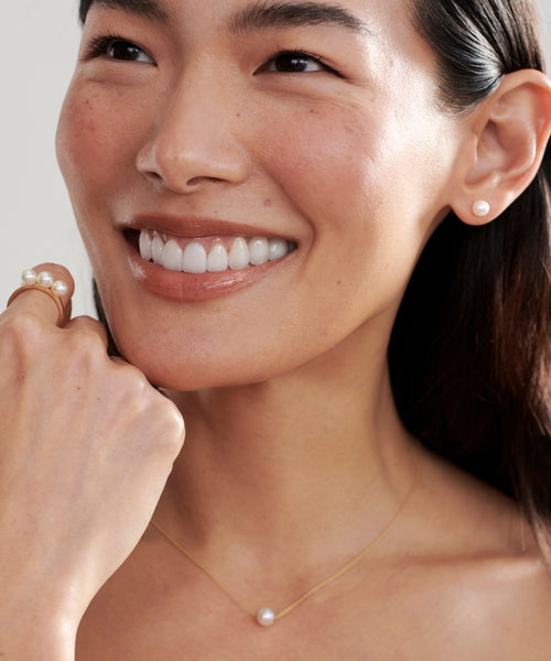 A woman smiles, showing white teeth. She wears pearl stud earrings, a delicate gold necklace with a single pearl, and the Elodie Pearl Ring—a 14k gold piece from our fine jewelry collection. Her hand touches her face, hair tucked behind one ear.