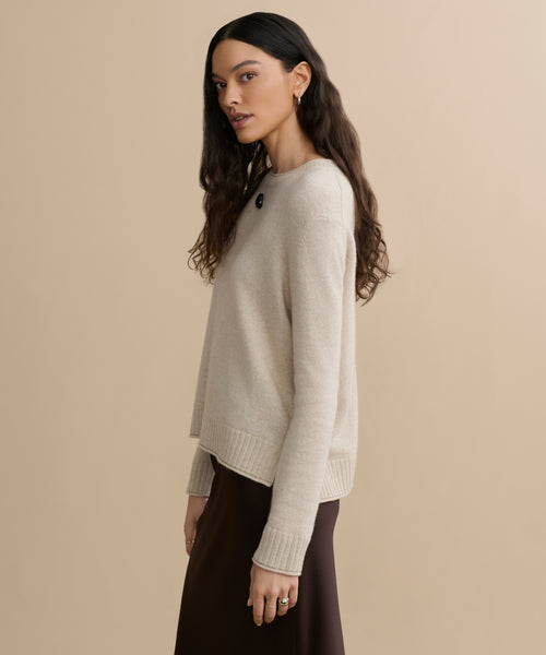 A woman with long, wavy dark hair wears the beige Everyday Sweater and a brown skirt, standing sideways against a plain beige background. She looks slightly toward the camera with a neutral expression.