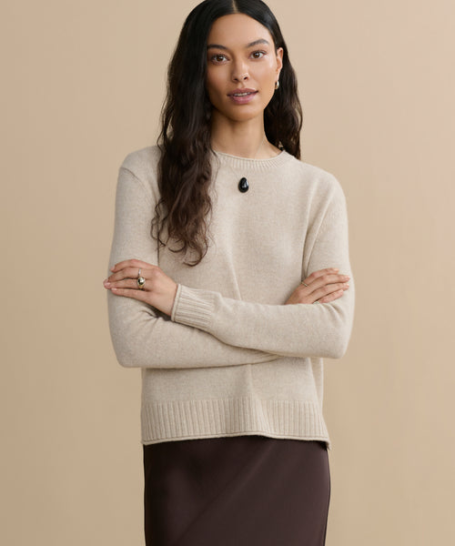 A woman with long dark hair stands against a beige background, wearing the Everyday Sweater, a dark brown skirt, a black pendant necklace, and a ring. She crosses her arms and gazes at the camera with a slight smile.