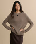 Woman with long, wavy dark hair stands against a beige background wearing the Everyday Sweater in brown, paired with a dark skirt and black pendant necklace, hands on her hips.