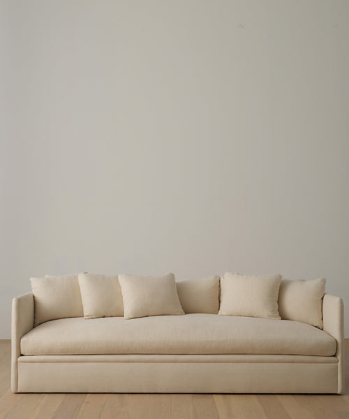 A modern sofa upholstered in Fabric Swatch Beige Hemp, accented with five matching cushions, sits against a plain light wall on a wooden floor.
