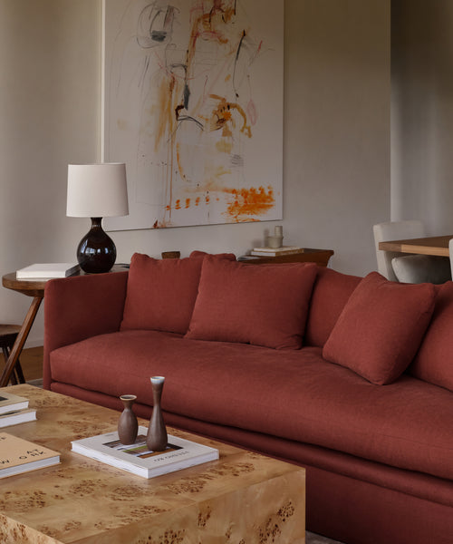 A modern living room with a Dume Sofa in Farmhouse Red Linen, a light wood coffee table with books and vases, a dark table lamp, and abstract artwork featuring orange accents above the sofa.